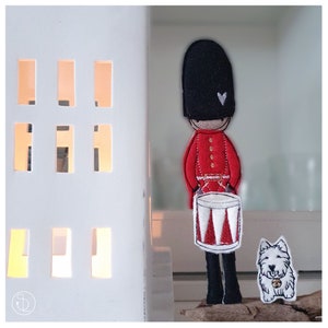 Könnte beinhalten: Ein Filzspielzeug eines britischen Wächters in roter Uniform, der eine Trommel hält, steht neben einem weißen Filzspielzeug-Hund mit einem roten Herzen auf der Brust.