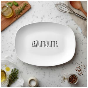 Op de afbeelding: Een witte, ovale serveerschaal met de tekst "KRÄUTERBUTTER" in zwarte letters. De afbeelding bevat kookingrediënten zoals kruiden, specerijen en olijfolie, wat wijst op een culinair thema.