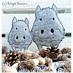 May include: Two gray felt wild boar figures with black embroidered details. The larger boar is standing behind the smaller boar. Both boars are smiling. The figures are sitting on a bed of pine cones and acorns.