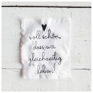 Könnte beinhalten: Weißer Stoff mit schwarzer Stickerei, die "voll schön, dass wir gleichzeitig leben!" lautet, mit einem kleinen schwarzen Herzen über dem Text.
