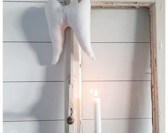 May include: A plush white tooth with a small gold heart detail hangs from a distressed white frame. A lit white candle in a black holder sits below. The background is a white paneled wall.