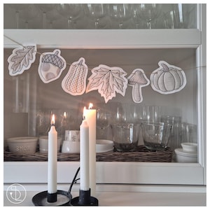 Puede incluir: Interior de un armario blanco con una guirnalda temática otoñal y velas encendidas. La guirnalda presenta formas de hojas, bellotas, calabazas y setas. Tres velas blancas en soportes negros están encendidas, creando un brillo cálido. Se ven cristalería y cuencos decorativos.