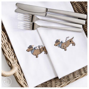 Könnte beinhalten: Zwei weiße Stoffservietten mit gestickten braunen Dackeln, die graue Hüte und blaue Halstücher tragen, belegt mit einem Edelstahlmesser und zwei Gabeln. Die Hunde haben kleine schwarze Herzmarkierungen an ihren Seiten und tragen Zubehör.
