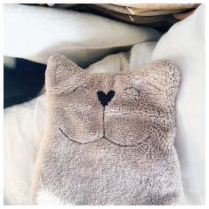 May include: A plush, light brown cat-shaped pillow with a black heart-shaped nose and embroidered smiling face. The pillow is resting on a white surface, creating a cozy and inviting aesthetic.