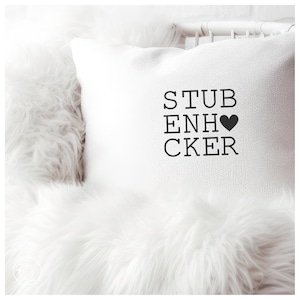 Könnte beinhalten: Weißes Dekokissen mit dem schwarzen Text "STUB ENH ❤️CKER". Das Kissen liegt auf weißem Kunstfell. Der Text enthält ein schwarzes Herzsymbol.