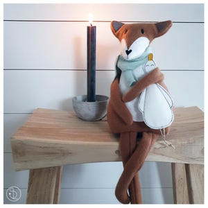 Può includere: Un peluche di volpe fatto a mano con una sciarpa verde chiaro, che tiene in braccio un uccello di peluche bianco. La volpe è marrone con la faccia bianca e le orecchie grigie. Una candela nera accesa è in un supporto di cemento su una panca di legno.