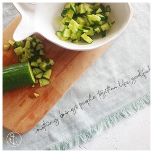 May include: A white bowl filled with chopped cucumbers on a wooden chopping board. A whole cucumber is next to the bowl. The text "nothing brings people together like good food" is written on a light blue fabric with a fringe border.