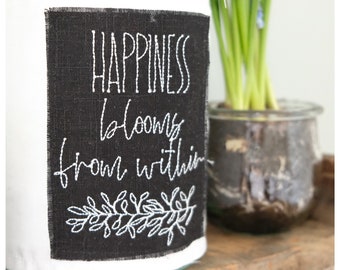 May include: A white glass vase with a black fabric label that says "Happiness blooms from within" in white embroidery. The vase is filled with green plant stems.