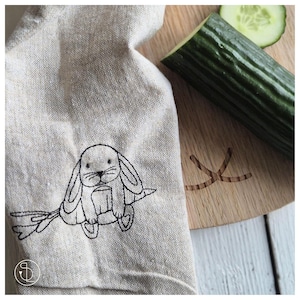 Könnte beinhalten: Ein beigefarbenes Leinen-Geschirrtuch mit einem schwarzen gestickten Design eines Kaninchens, das eine Karotte hält. Das Kaninchen sitzt auf einem Ast mit Blättern.