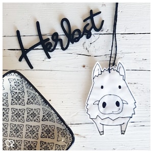 May include: A white fabric ornament with black embroidery of a wild boar. The ornament is hanging from a black string. The word "Herbst" is written in black on a white wooden background.
