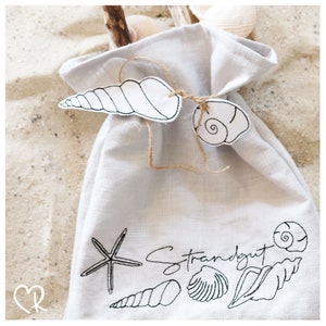 Könnte beinhalten: Eine weiße Leinentasche mit Kordelzug. Die Tasche ist mit schwarzen gestickten Muscheln, einem Seestern und dem Text "Strandgut" verziert.