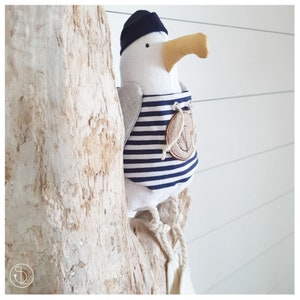 Peut inclure: Un goéland en peluche blanc portant une chemise rayée bleu et blanche avec une ancre sur le devant. Le goéland porte un chapeau de marin bleu et est perché sur un morceau de bois flotté.