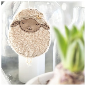 Puede incluir: Adorno de oveja color crema con una cara sonriente, detalles cosidos y un pequeño botón de madera. La oveja tiene un cuerpo esponjoso y texturizado, suspendido por una cuerda. Una decoración encantadora y artesanal.