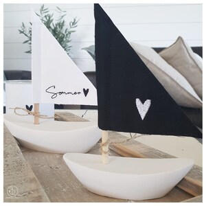 May include: Two white ceramic sailboats with wooden masts. One sailboat has a white sail with the word "Sommer" and a black heart. The other sailboat has a black sail with a white heart.
