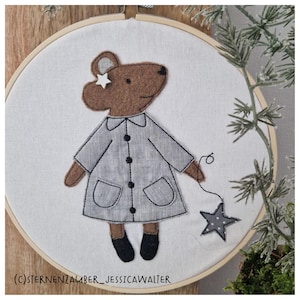 May include: Embroidered brown bear wearing a grey coat with black buttons and holding a star. The bear is in a white embroidery hoop.