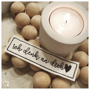 May include: A white ceramic candle holder with a lit tea light candle. The candle holder is surrounded by wooden beads. A white fabric tag with black embroidery reads "ich denk an dich" with a small black heart.