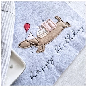 May include: A light blue felt fabric with a brown dachshund wearing a party hat and carrying a pink gift box and a cake. The text "happy birthday" is embroidered in black thread.