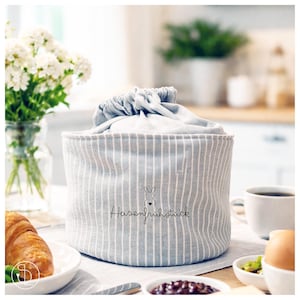 Puede incluir: Una cesta de tela a rayas grises y blancas con cierre de cordón, con una ilustración de conejo y el texto "Hasenfrühstück". La cesta está sobre una mesa de desayuno, con un cruasán y café.