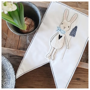 Op de afbeelding: Een witte stoffen banner met een handgestikte geborduurde konijn dat een bloem vasthoudt. Het konijn is beige met een blauwe strik en een zwart hart. De bloem is paars.