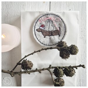 Könnte beinhalten: Ein runder, grauer Weihnachtsbaumschmuck aus Filz mit einem Dackel, der einen Weihnachtsmannhut trägt und die Zahl 13. Der Schmuck ist mit rot-weißem Garn verziert. Eine brennende Kerze und ein Zweig mit Tannenzapfen sind ebenfalls im Bild.