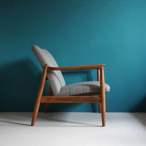 May include: A gray upholstered armchair with a wooden frame. The chair has a simple, mid-century modern design and is set against a teal wall.