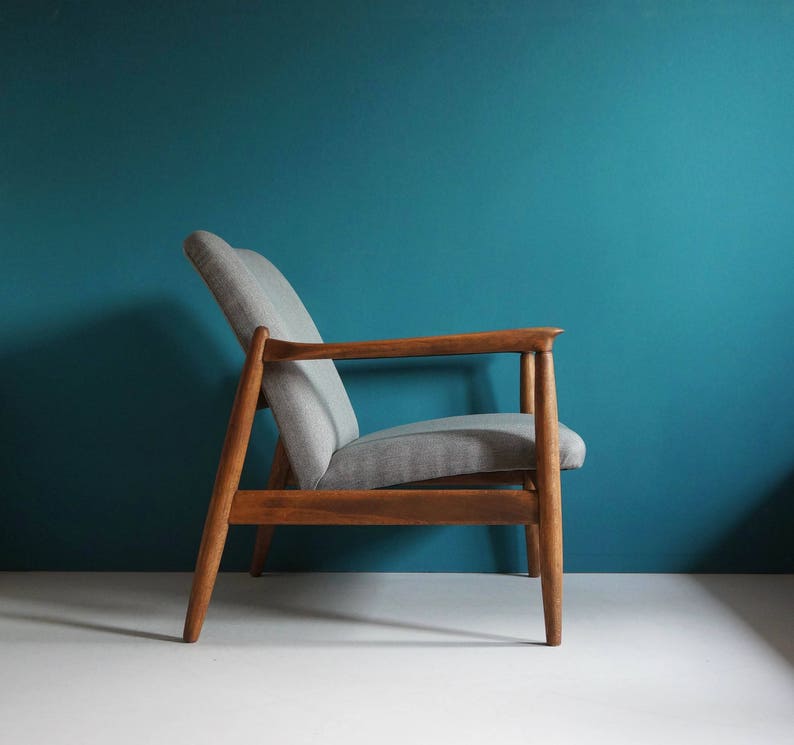 May include: A gray upholstered armchair with a wooden frame. The chair has a simple, mid-century modern design and is set against a teal wall.