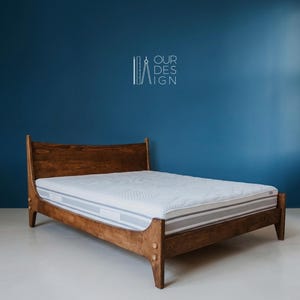 Op de afbeelding: Een donkerbruin houten bedframe met een gebogen hoofdeinde en vier poten, voorzien van een wit matras met een doorgestikt bovenoppervlak. De tekst "OUR DESIGN" is boven het bed zichtbaar, samen met een icoon van een liniaal en passer.