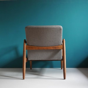 May include: A gray upholstered armchair with a wooden frame. The chair has a simple design with a high back and a low seat. The chair is sitting on a white surface in front of a teal wall.