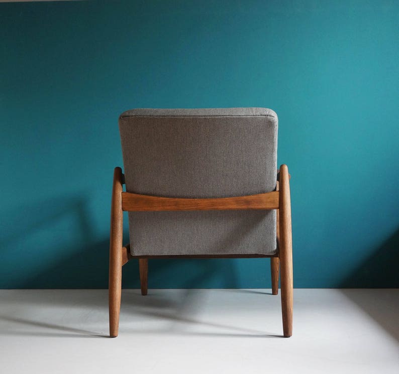 May include: A gray upholstered armchair with a wooden frame. The chair has a simple design with a high back and a low seat. The chair is sitting on a white surface in front of a teal wall.