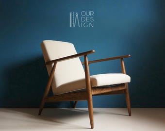 Può includere: Una poltrona bianca con braccioli e gambe in legno. La poltrona è inclinata a sinistra e lo sfondo è una parete blu tinta unita. Il testo "OUR DESIGN" è visibile nell'angolo in alto a destra dell'immagine.