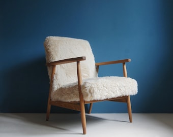 May include: A white faux fur armchair with a wooden frame. The chair has a simple design with straight lines and a low back.