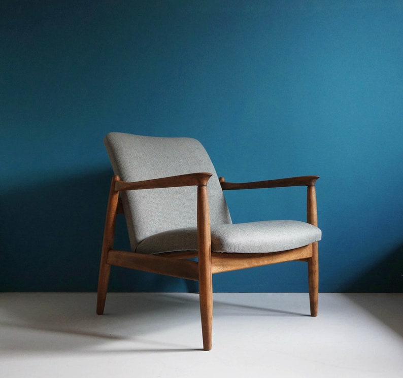 May include: A light gray armchair with a wooden frame. The chair has a simple design with a high back and armrests. The chair is sitting on a light blue floor.