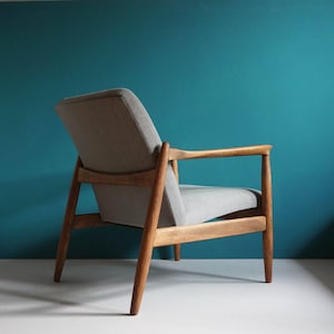 May include: A gray upholstered armchair with a wooden frame. The chair has a simple design with a low back and wide arms. The chair is sitting on a teal blue background.