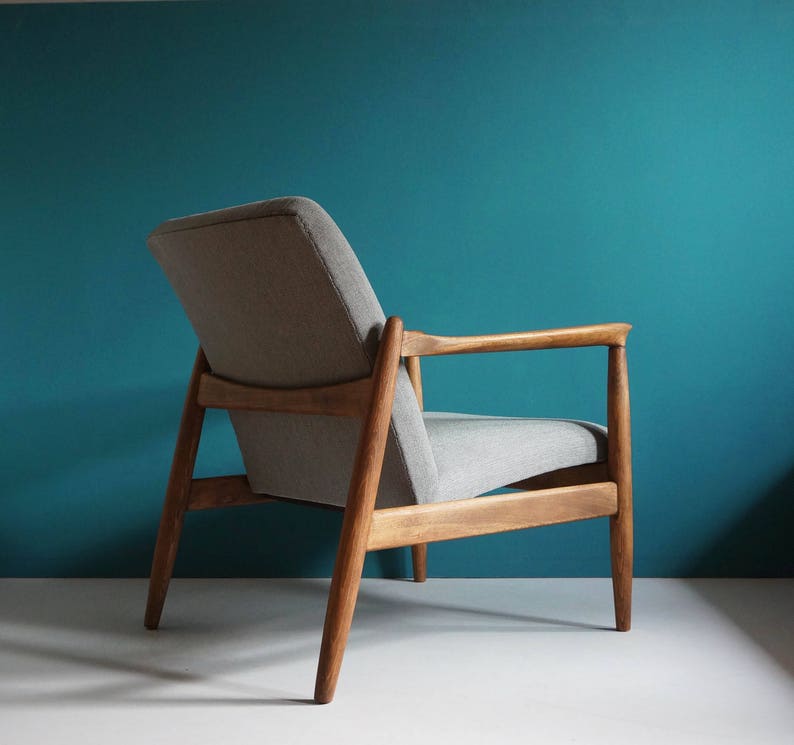 May include: A gray upholstered armchair with a wooden frame. The chair has a simple design with a low back and wide arms. The chair is sitting on a teal blue background.