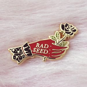 Può includere: Spilla in smalto dorato raffigurante una mano rossa che tiene una rosa nera. La mano ha le parole "BAD SEED" in bianco. Il polsino è nero con un disegno di stelle e luna.