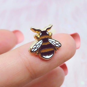 Puede incluir: Pin de esmalte de abeja dorada con rayas negras y amarillas. La abeja tiene alas pequeñas y un cuerpo detallado.