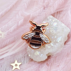 May include: A gold-tone bee pin with black and white stripes and clear enamel accents. The pin is sitting on a white crystal cluster.