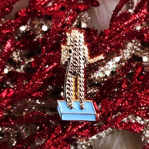 May include: Gold-colored enamel pin featuring a cartoon figure standing on a blue platform. The figure is wearing a long, shaggy coat and has a long, thin nose.
