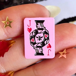 Op de afbeelding: Een roze speelkaartsieraden met de boer van harten, met zwart-witte details en rode hartsymbolen. Gouden stervormige versieringen en rode bloemblaadjes omringen de kaart.