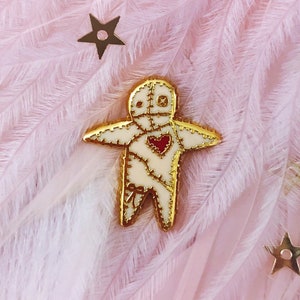 May include: Gold-tone pin featuring a white voodoo doll with a red heart and stitched details. The pin is on a pink feather background.