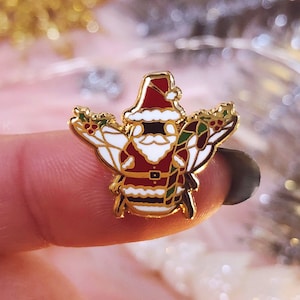 May include: Gold-tone enamel pin featuring a cartoon Santa Claus dressed as a bee, holding a candy cane and holly.