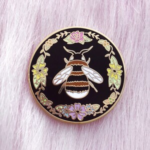 May include: A gold-toned enamel pin featuring a black and yellow bumblebee surrounded by a wreath of colorful flowers on a black background.