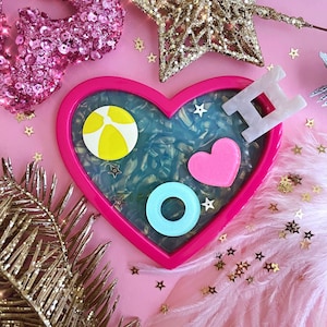May include: A pink heart-shaped tray with a blue and white marbled design. The tray features a yellow beach ball, a blue and white ring, a pink heart, and a white letter H.