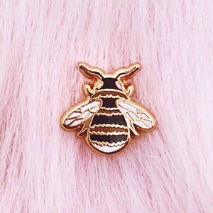 May include: A gold-toned bee pin with black and white stripes and white wings. The pin is on a pink fuzzy background.