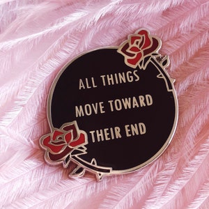 May include: Black enamel pin with a gold border and two red roses. The text on the pin reads "All things move toward their end."