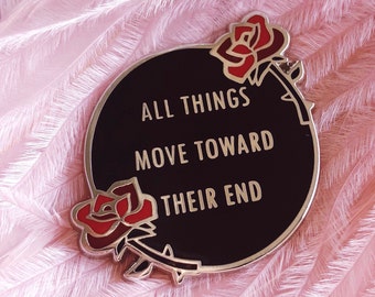 May include: Black enamel pin with a gold border and two red roses. The text on the pin reads "All things move toward their end."