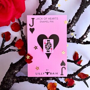K&ouml;nnte beinhalten: Eine rosa Karte mit einem schwarzen Herz und dem Text "JACK OF HEARTS ENAMEL PIN" zeigt eine kleine Anstecknadel einer Spielkarte. Die Karte ist mit goldenem sternf&ouml;rmigem Konfetti und roten Rosenbl&auml;ttern verziert. Die Karte wurde von Lilly Baik entworfen.
