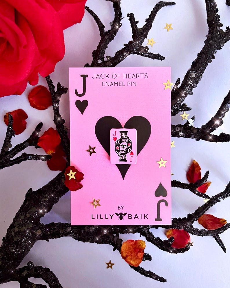 K&ouml;nnte beinhalten: Eine rosa Karte mit einem schwarzen Herz und dem Text "JACK OF HEARTS ENAMEL PIN" zeigt eine kleine Anstecknadel einer Spielkarte. Die Karte ist mit goldenem sternf&ouml;rmigem Konfetti und roten Rosenbl&auml;ttern verziert. Die Karte wurde von Lilly Baik entworfen.