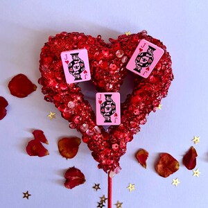 K&ouml;nnte beinhalten: Rote, herzf&ouml;rmige Dekoration mit Pailletten, mit drei rosa Spielkarten mit einem schwarz-wei&szlig;en Design. Rosenbl&auml;tter und goldfarbene Sternkonfetti sind verstreut.