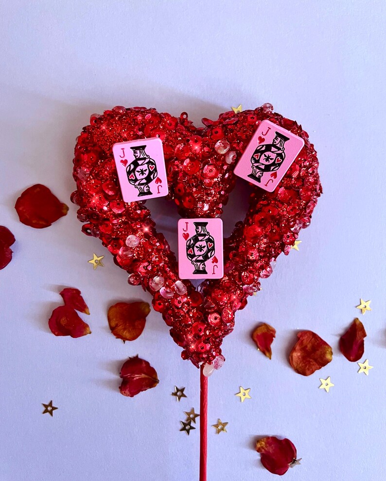 K&ouml;nnte beinhalten: Rote, herzf&ouml;rmige Dekoration mit Pailletten, mit drei rosa Spielkarten mit einem schwarz-wei&szlig;en Design. Rosenbl&auml;tter und goldfarbene Sternkonfetti sind verstreut.